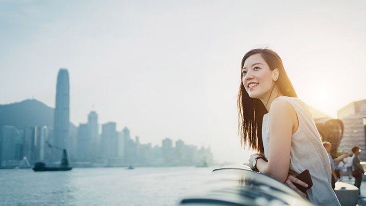 Smiling young Asian woman traveller looking out over promenade of Victoria Harbour enjoying the gentle breeze and the beautiful cityscape at dusk