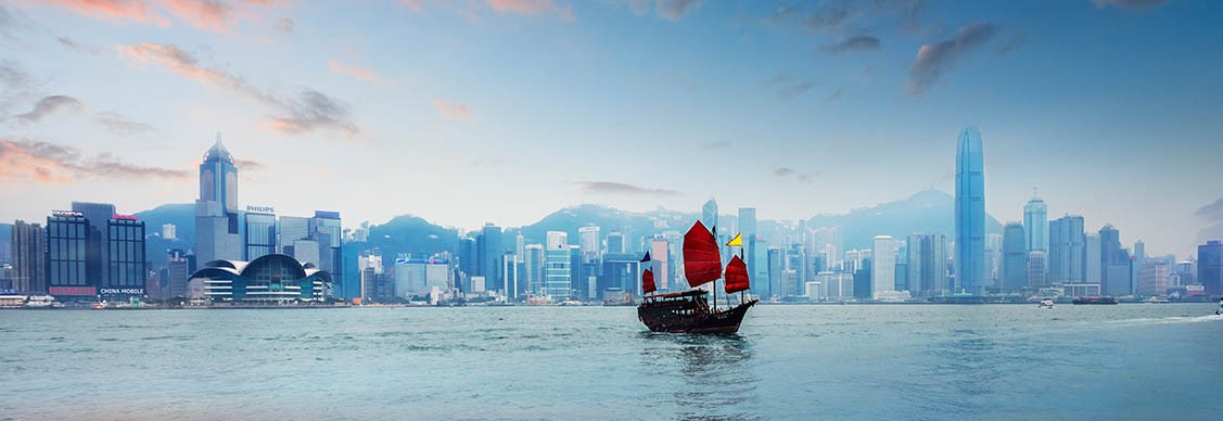 A colorful boat with red sails sails smoothly in the water, with a cityscape visible in the distance.