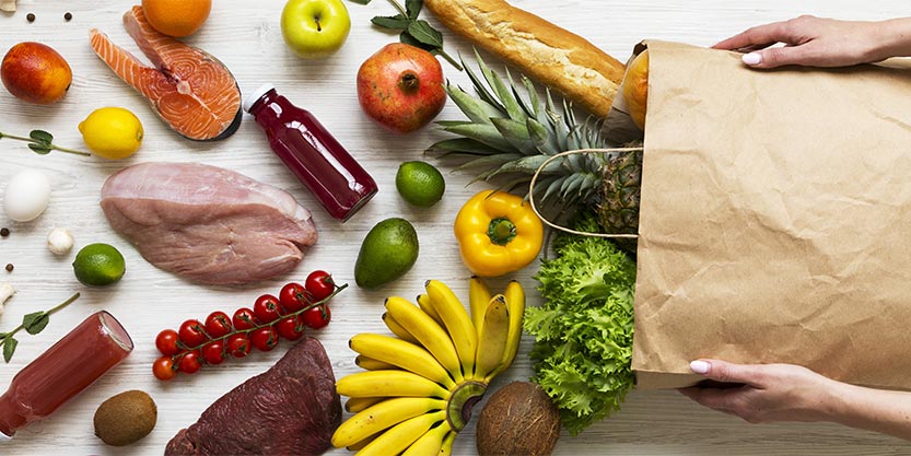 Women's hands hold full paper bag of groceries over white wooden background, top view. From above, overhead. Flat lay.