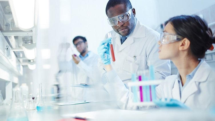 Female Asian laboratory scientist in lab coat and safety goggles showing test tube with red liquid to curious African-American colleague in laboratory. Latin-American scientist in background.