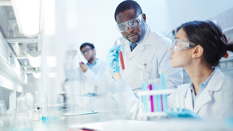 Female Asian laboratory scientist in lab coat and safety goggles showing test tube with red liquid to curious African-American colleague in laboratory. Latin-American scientist in background.