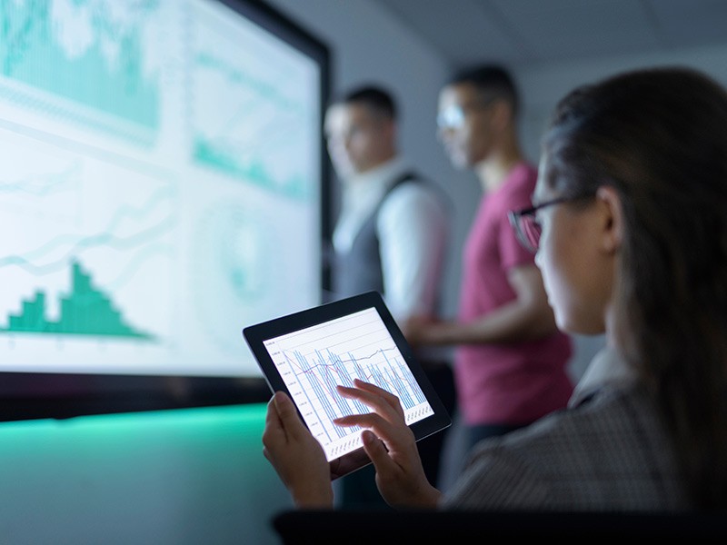 Businesswoman reviews data and charts on a digital tablet in a business meeting, while co-workers view data on a larger screen in the background