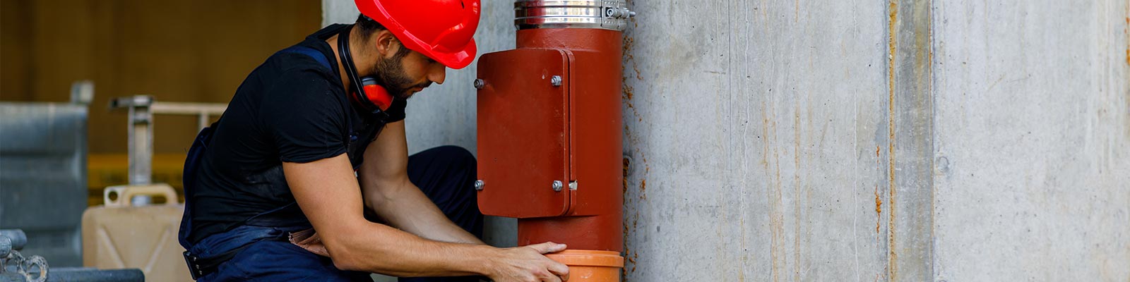 A facilities management professional in hard hat repairs a large pipe in an industrial setting